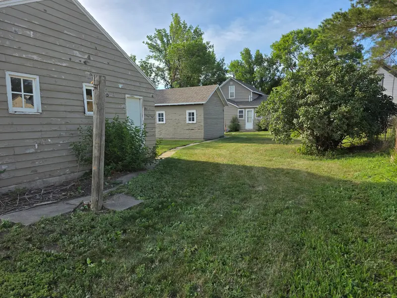 A mown backyard beside a grey house, with a large bush and an outbuilding behind