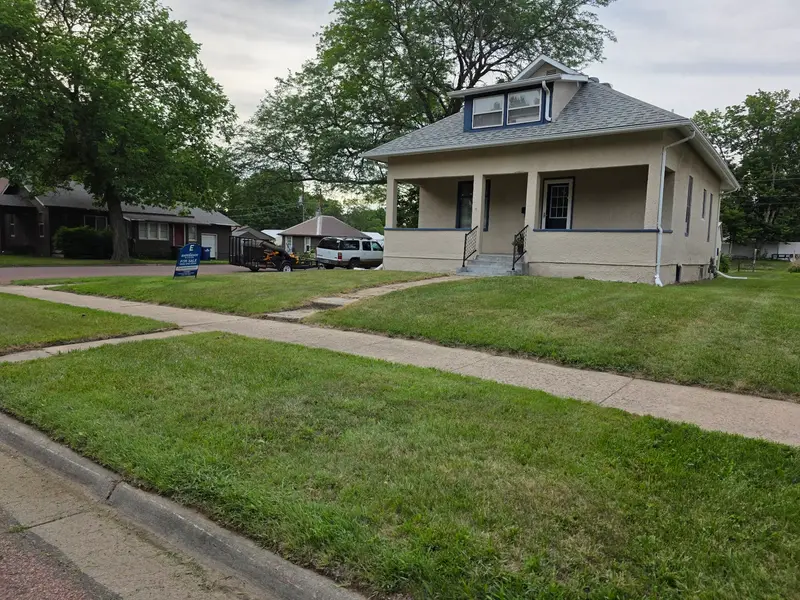 A mown front lawn and sidewalk strip in front of a tan bungalow with a covered porch